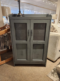 The 357 Punched Tin Cupboard by Woodcraft is an Amish-made, gray wooden cabinet featuring decorative star-patterned punched tin panels on the doors, providing ample storage. Showcased beautifully in a furniture store, this piece blends rustic charm with practical design.