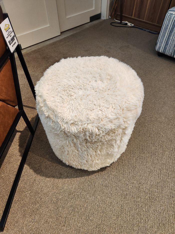 A cozy, fluffy 77004 Mingle Small Round Ottoman by Palliser with single top stitching sits on a carpeted floor, beckoning as the perfect furniture upgrade, its price tag displayed nearby.