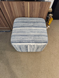 Palliser's 77004 Mingle Small Square Ottoman with single top stitching on a beige carpeted floor, near wooden furniture.