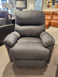 The furniture showroom features the 7MP84 Ellisport Power Space Saver Recliner from Best, showcased in a luxurious gray finish that promises unparalleled comfort. Its plush back and seat invite relaxation, complemented by elegant sofas and ambient lamps in the background.