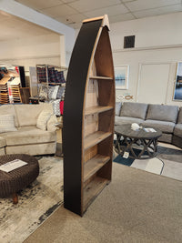 The 322 7' Boat Bookcase by Woodcraft is creatively showcased in a furniture showroom, highlighting smart storage solutions. Flanked by sofas and decor, these shelves exemplify effective merchandising while adding a nautical charm to the space.