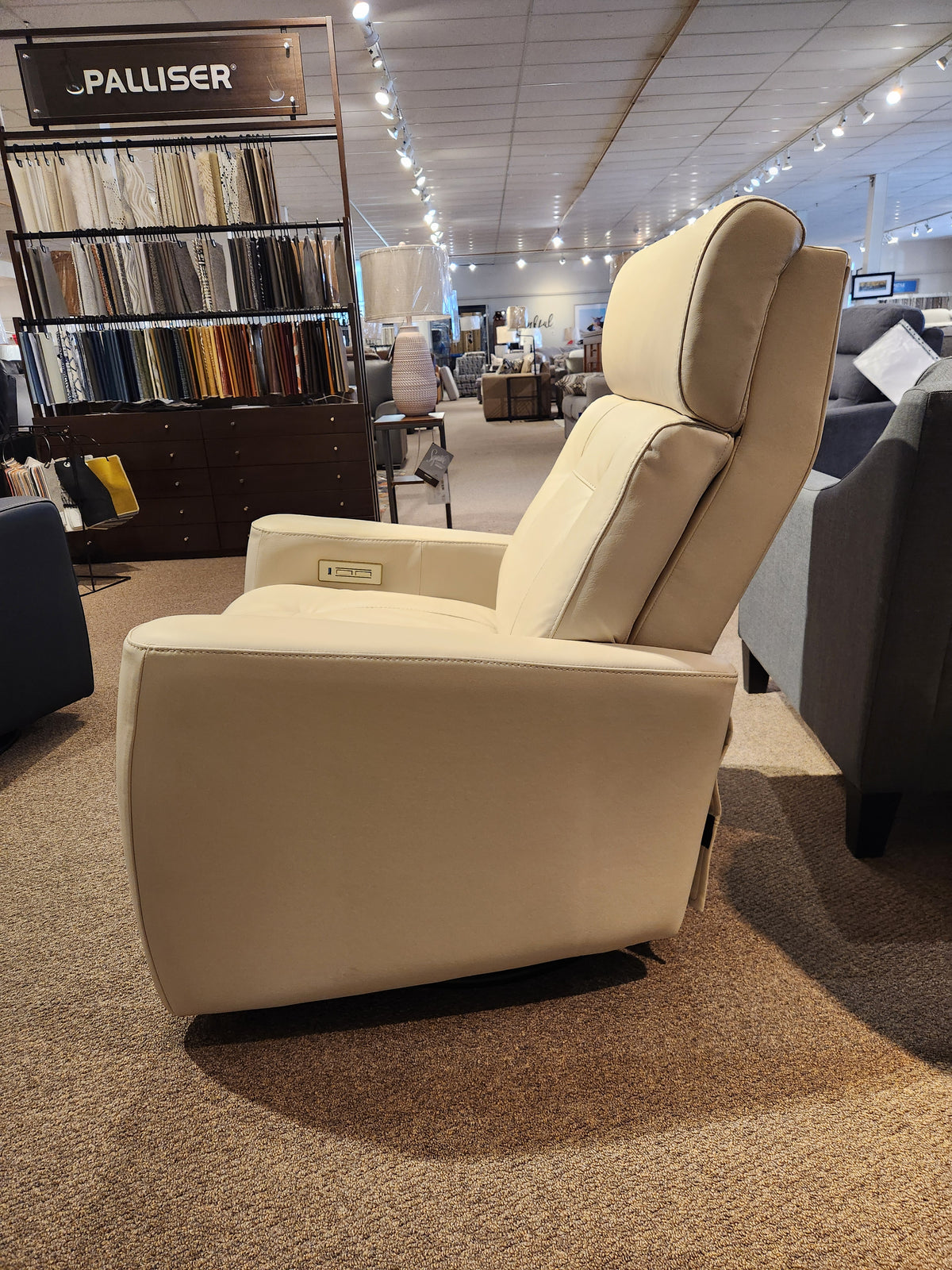 The Palliser 42204 West Coast Power Swivel Glider, a beige recliner, sits in a showroom with fabric samples and other seating.