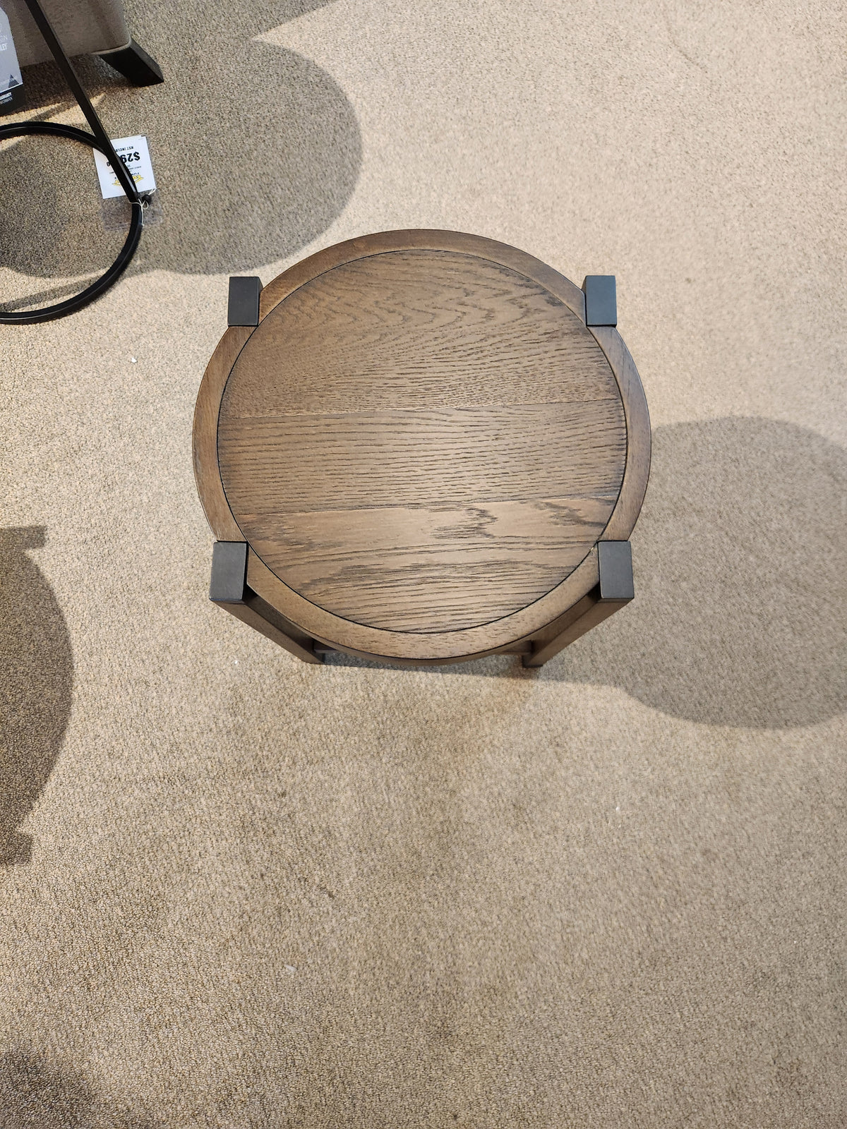 Top view of the T5817-05 Kaysen Round End Table by Magnussen, featuring dark metal accents, set on a beige carpeted floor, complementing the Wallaby finish of the Modern Organic Collection.