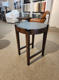 The T5817-05 Kaysen Round End Table from Magnussen's Modern Organic Collection, featuring black accents, sits elegantly in a showroom. It complements the chairs set on the carpeted floor and fabric samples nearby. Its sleek Wallaby finish mirrors the refined elegance of the nesting tables displayed around it.