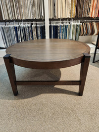 Magnussen's T5817-45 Kaysen Round Cocktail Table, part of the Modern Organic Collection, rests on a carpeted floor. It features matte black hardware and is surrounded by hanging fabric samples in a store setting.