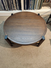 The Magnussen Kaysen Round Cocktail Table, model T5817-45, from the Modern Organic Collection, elegantly stands on a carpeted floor with its dark-finished wooden surface and Matte Black hardware accentuating the corners.