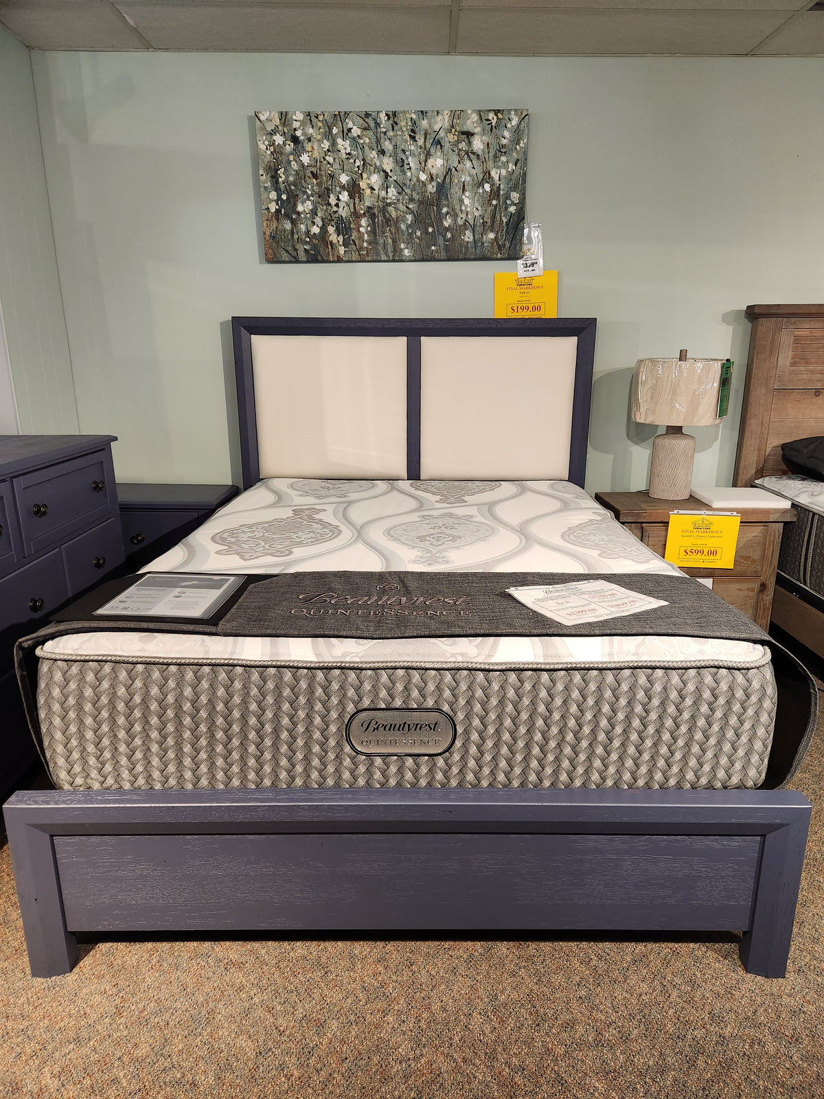 Bedroom display featuring a Vaughan Bassett upholstered bed, patterned mattress, navy frame, bedside lamp, and abstract wall art.