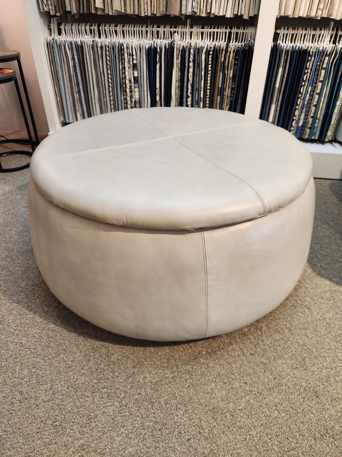 An Oliver Round Storage Ottoman from La-z-boy, featuring a beige leather design, is ideal for extra seating and is placed on a carpeted floor before a shelf brimming with various fabric samples.