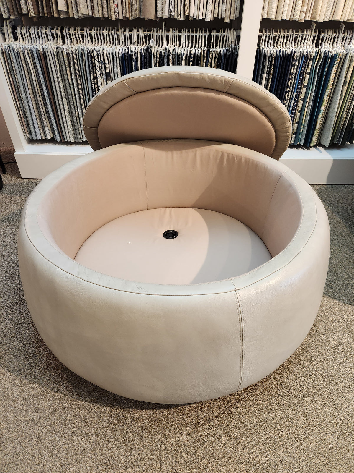 The Oliver Round Storage Ottoman by La-z-boy is an elegant beige leather piece featuring an open lid for storage, doubling as extra seating. Its circular design is showcased on a carpeted floor amid fabric samples.