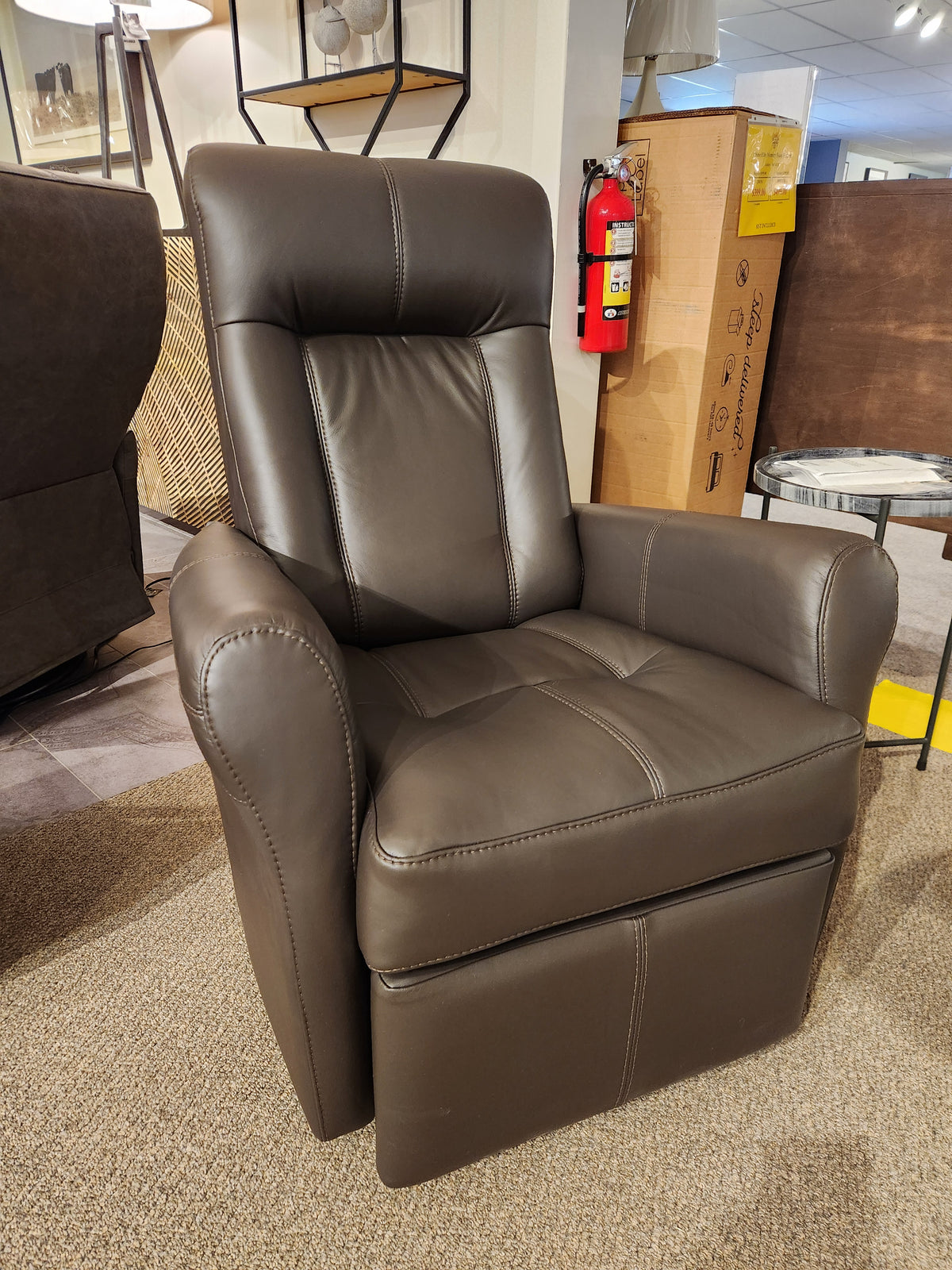 The showroom displays the 42201 Yellowstone I Rocker Recliner by Palliser, offering ergonomic comfort with its brown leather design. In the background, a fire extinguisher and cardboard boxes complete the scene.