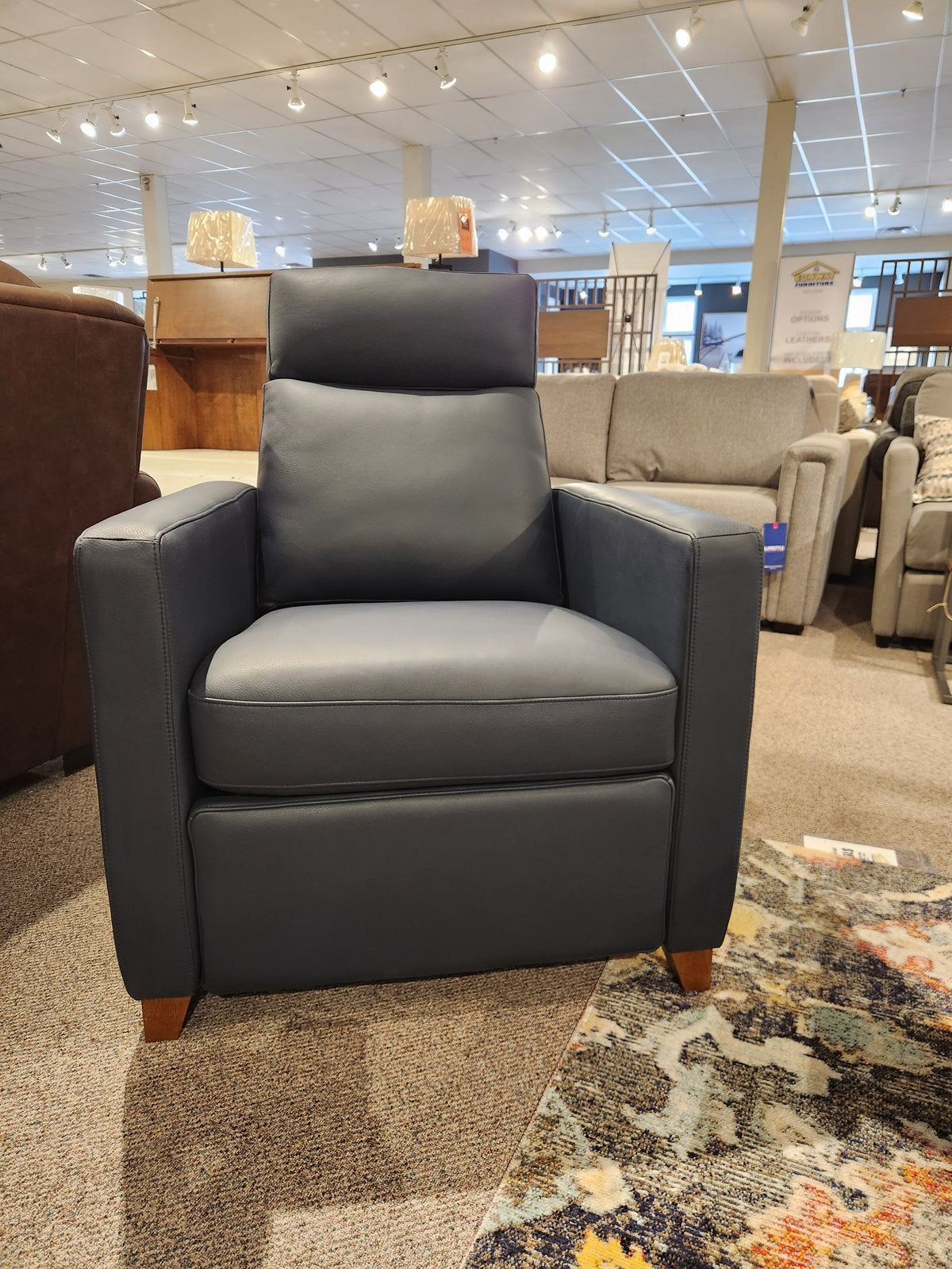 The Palliser 77171 Soren Push-Back Reclining Chair in dark gray leather, showcasing classic styling with modern Soren upholstery, is displayed in a furniture showroom, highlighting its elegant tapered wooden legs that perfectly blend traditional and contemporary design.