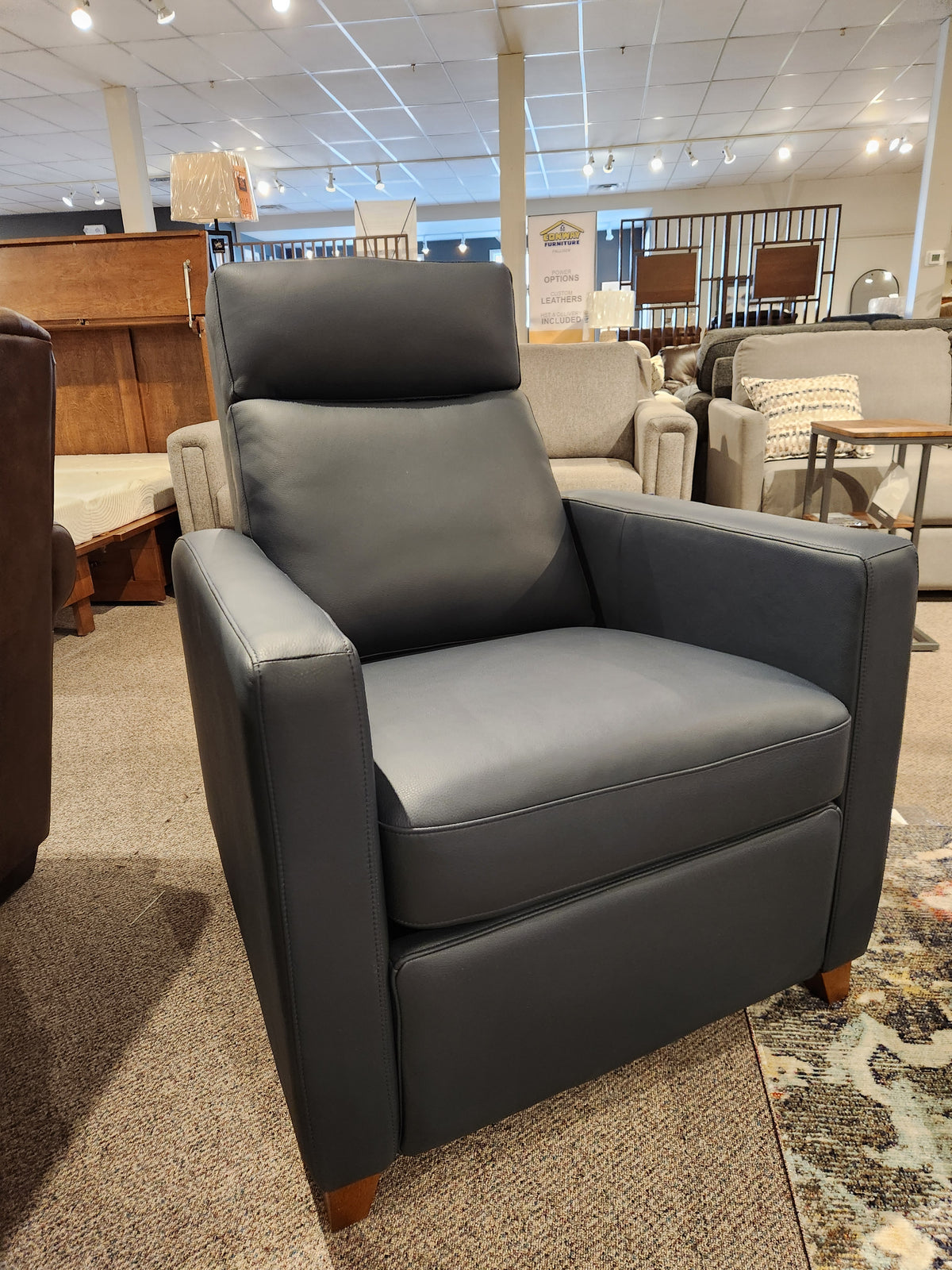 A stylish leather armchair, the 77171 Soren Push-Back Reclining Chair by Palliser, featuring tapered wooden legs and classic design, is showcased in a furniture store surrounded by other chairs and tables on a carpeted floor.