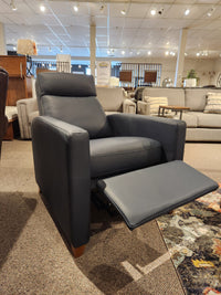 A 77171 Soren Push-Back Reclining Chair by Palliser, with a gray Soren upholstery and extended footrest, is displayed in a furniture showroom. The classic styling is complemented by surrounding sofas, decor items, and tapered wooden legs.