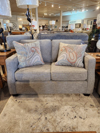 The Decor-rest 2570 Stationary Loveseat, crafted in Canada and featuring a stylish gray design with patterned pillows, is ideal for small spaces and is elegantly displayed on a beige rug in the store.