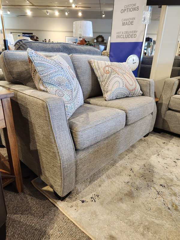 The Decor-rest 2570 Stationary Loveseat, featuring two patterned pillows, sits elegantly on a beige rug in the showroom, ideally suited for small spaces. A sign in the background emphasizes Canadian-made custom options.