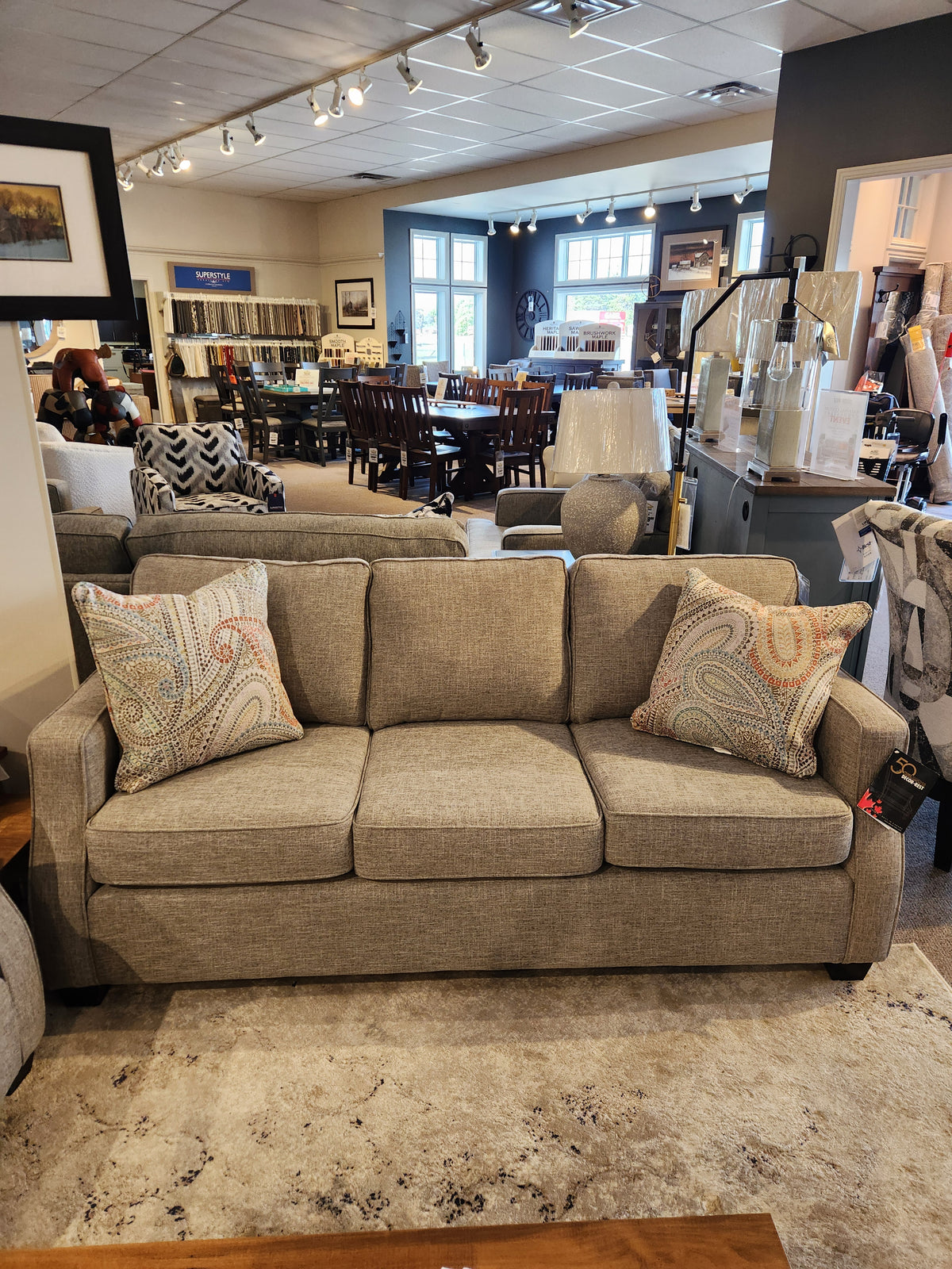 A 2570 Stationary Sofa by Decor-rest, featuring gray upholstery and patterned pillows, is showcased in a furniture store showroom. Made in Canada and designed for compact areas, it is surrounded by an assortment of tables and chairs.