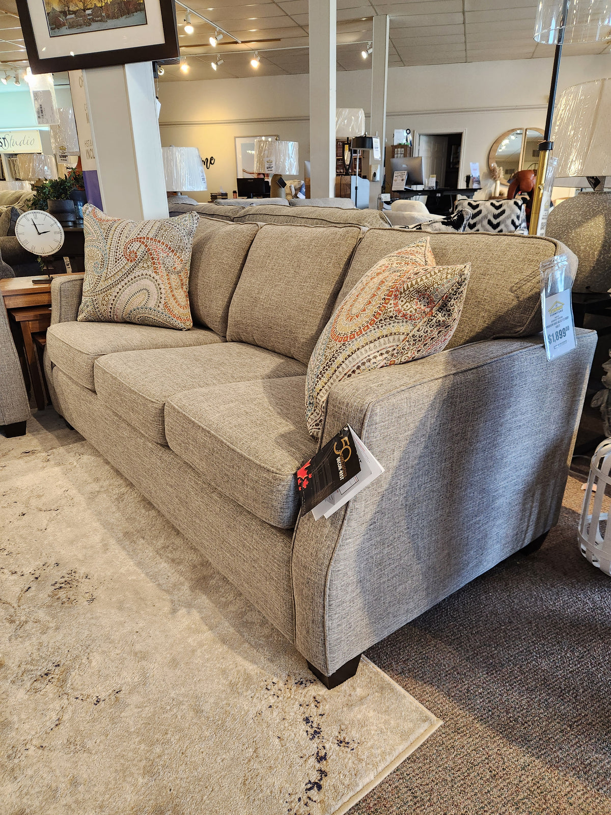 The Decor-rest 2570 Stationary Sofa, adorned with patterned throw pillows, offers an ideal solution for small spaces and is elegantly showcased in a furniture store showroom. This stylish piece, proudly Canadian-made, has a price tag attached to the arm.