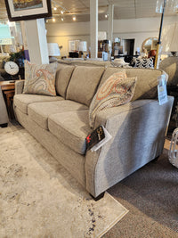 The Decor-rest 2570 Stationary Sofa, adorned with patterned throw pillows, offers an ideal solution for small spaces and is elegantly showcased in a furniture store showroom. This stylish piece, proudly Canadian-made, has a price tag attached to the arm.