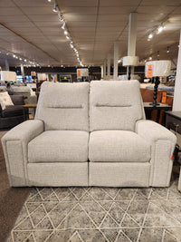 Displayed in a well-lit furniture store showroom is the M8500 Power Reclining Loveseat by Decor-rest, featuring elegant upholstery in light gray. This Canadian-made loveseat showcases its quality amidst various lamps and other furniture pieces in the background.