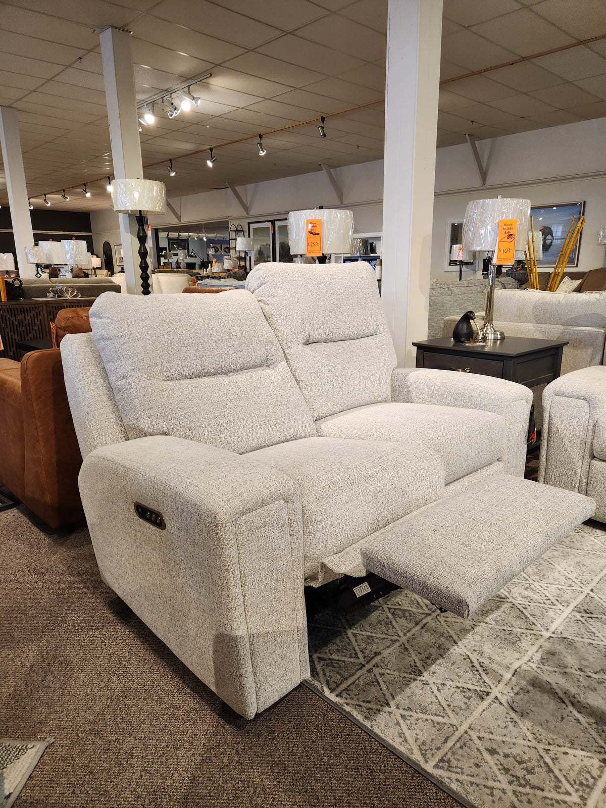 A Decor-rest M8500 Power Reclining Loveseat, upholstered in quality beige fabric, is showcased in the furniture showroom. Its adjustable footrests are extended, surrounded by other Canadian-made pieces.