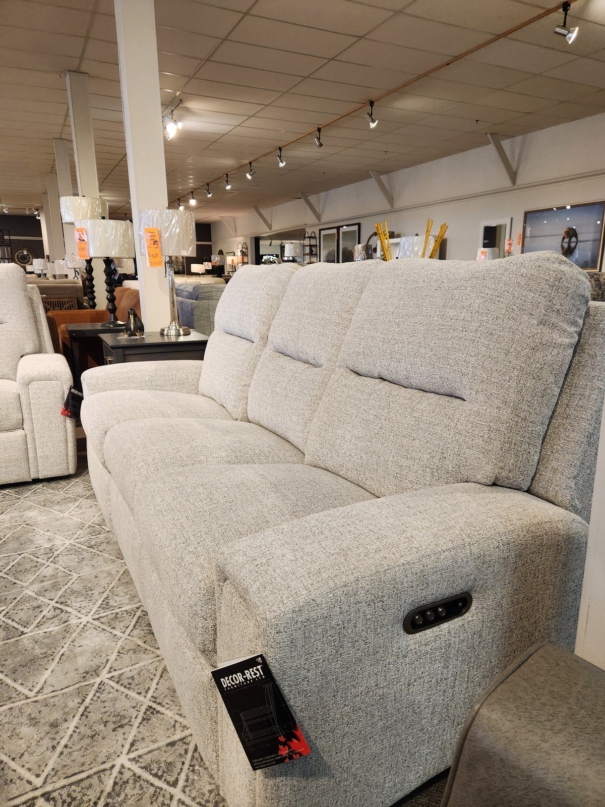 The M8500 Power Reclining Sofa by Decor-rest, featuring premium Canadian upholstery in a sophisticated gray, is showcased in a furniture store. It sits atop a geometric-patterned rug and is accented by elegant lamps and chic decor in the background.