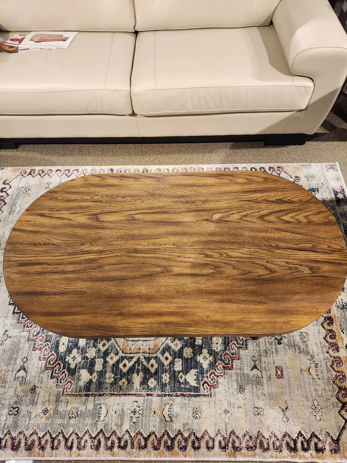 The Austanny T683 Coffee Table by Ashley, featuring a rustic oak veneer and an oval wooden design, sits gracefully on a decorative rug, complementing the beige couch.