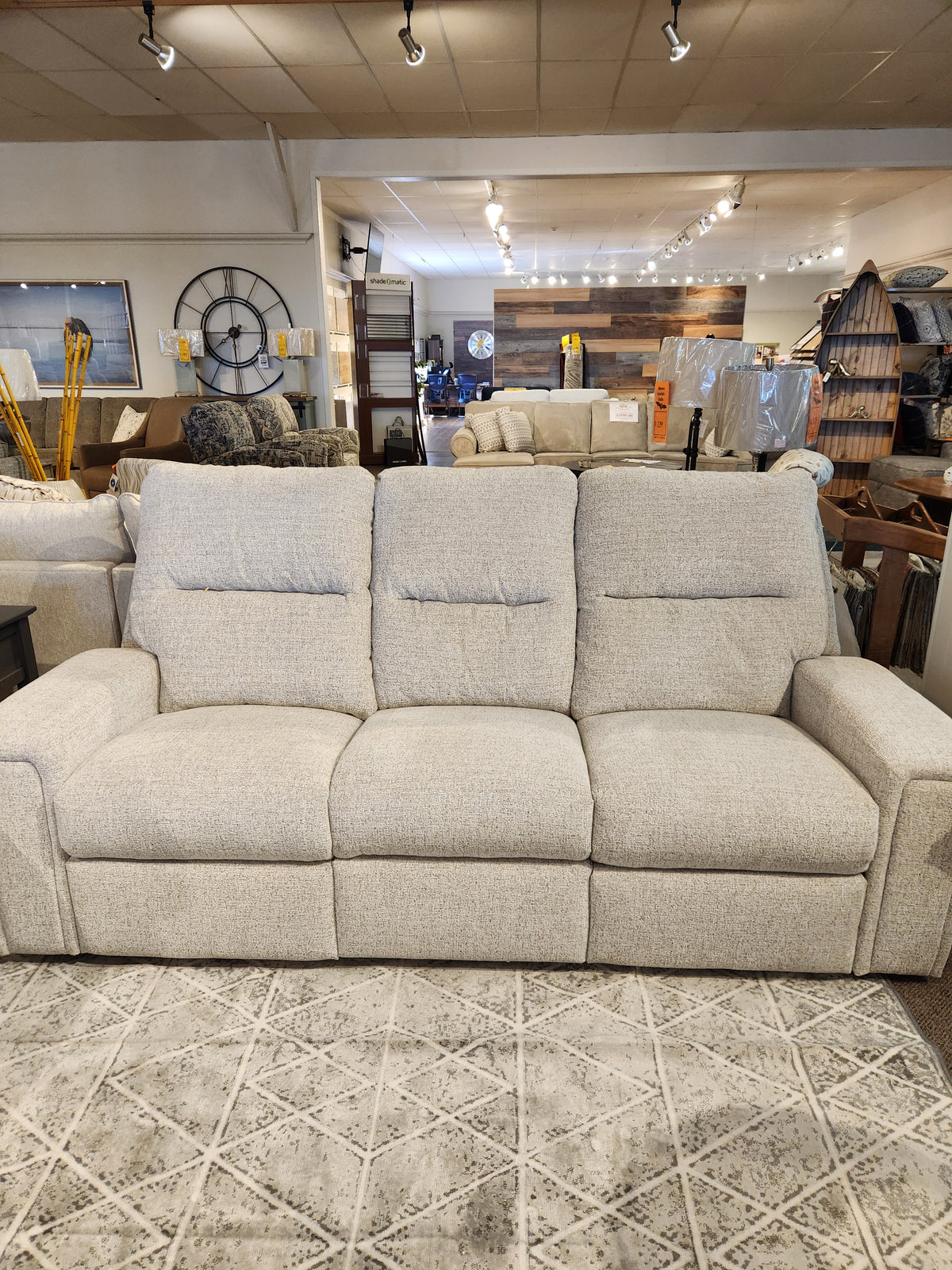 A Decor-rest M8500 Power Reclining Sofa, featuring a plush beige three-seater design, is elegantly displayed in the showroom, showcasing its exquisite upholstery amid various decor items in the background.