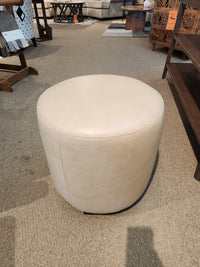 The Palliser 77004 Mingle Small Round Ottoman, featuring single top stitching, sits elegantly on a carpeted floor in a furniture store display, offering a chic furniture upgrade.