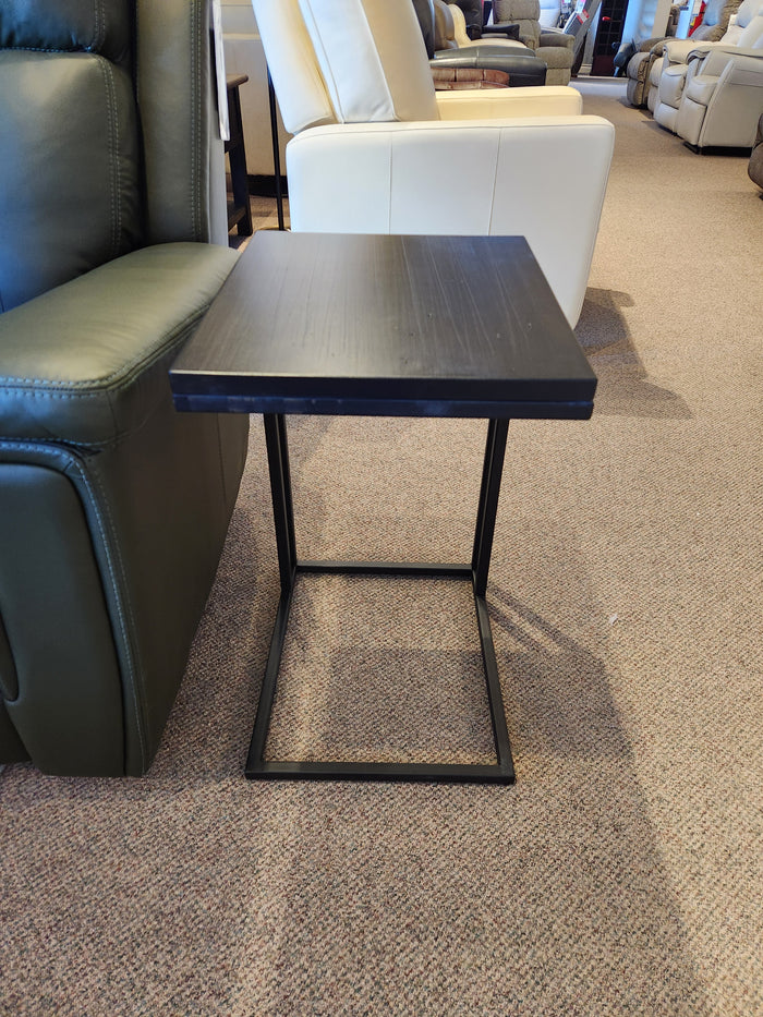 The Handstone AlaCarte Snack Table, a black metal and wood accent, is next to a green armchair on a carpeted showroom floor.