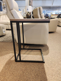 The AlaCarte Snack Table by Handstone, featuring a wooden top and black metal frame, stands on a showroom's carpeted floor.