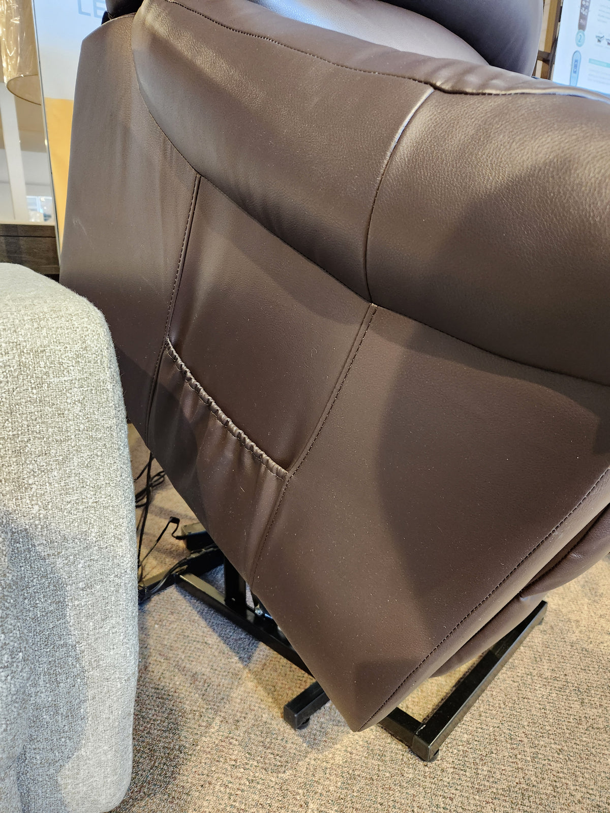 Primo's Fresna Power Lift Chair in brown faux leather with a side pocket, beside a gray fabric couch on carpet.