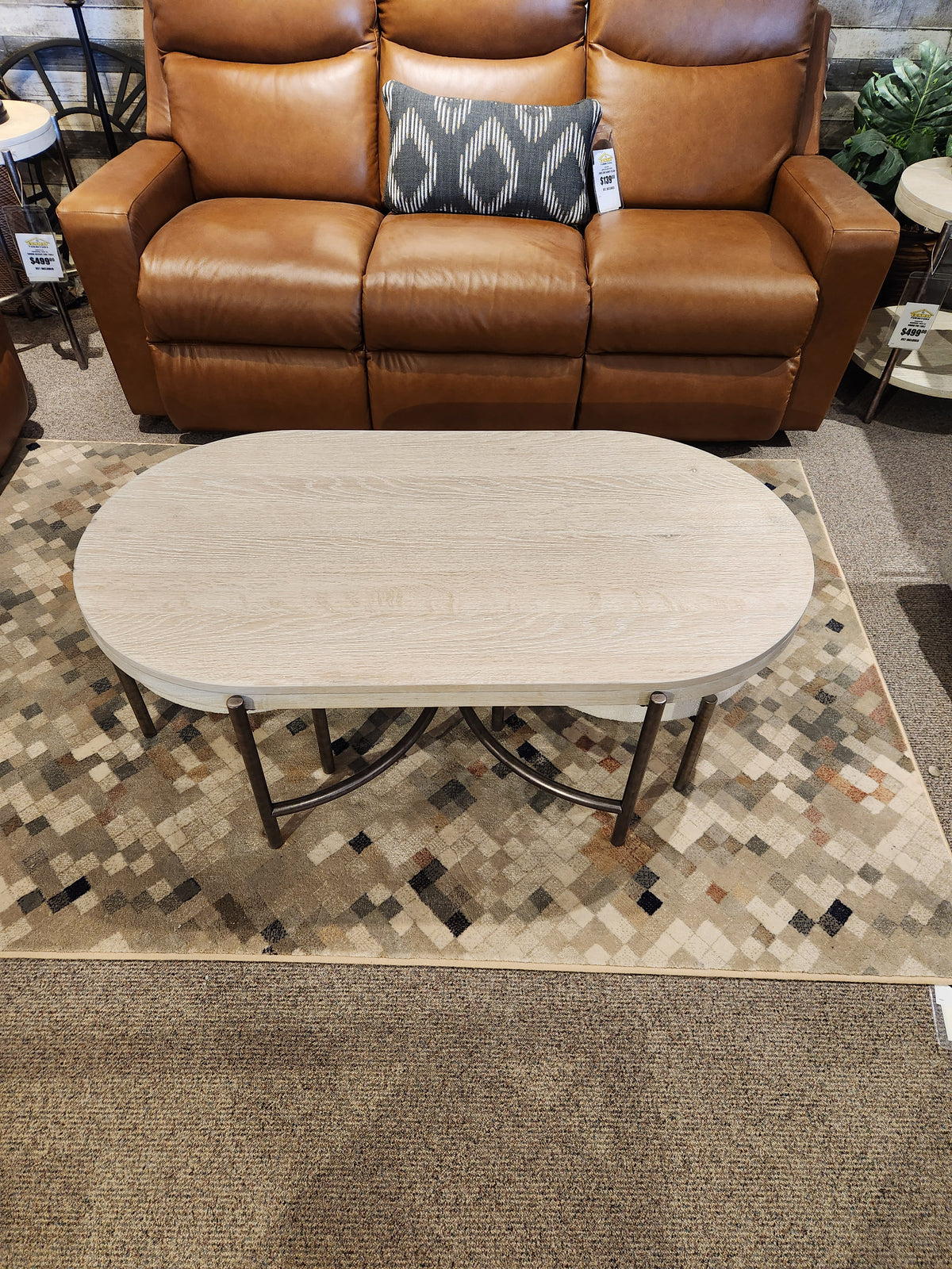 The Magnussen T5927 Jennings Oval Coffee Table in Edgecomb Grey sits on a patterned rug before a brown leather sofa.