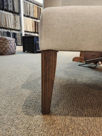 A beige Belmont Wing Back Chair by Handstone with oak legs on a carpeted floor in a fabric showroom.