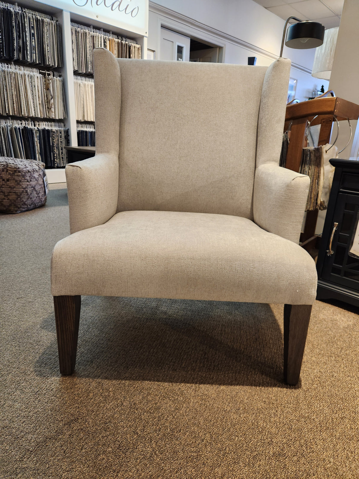 The Belmont Wing Back Chair by Handstone, with sturdy oak legs, stands gracefully on a carpeted showroom floor.