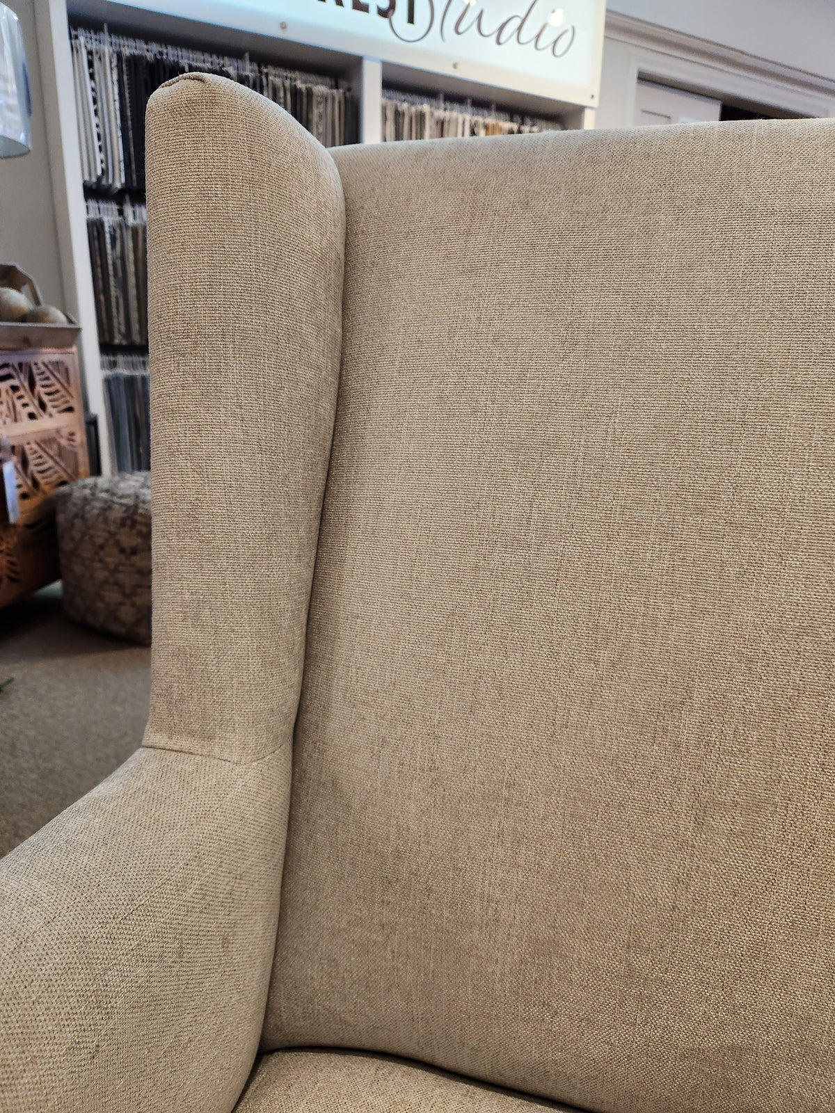 Close-up of the Handstone Belmont Wing Back Chair with a beige design and oak legs against a cluttered fabric store backdrop.