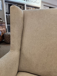 Close-up of the Handstone Belmont Wing Back Chair with a beige design and oak legs against a cluttered fabric store backdrop.