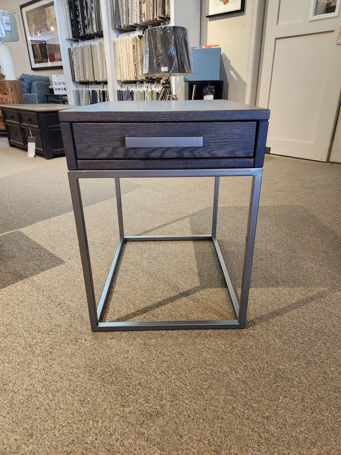 A solid oak Muskoka 18" End Table with a drawer and metal legs by Handstone on a carpeted showroom floor.