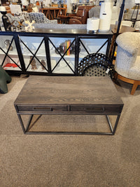 A Handstone Muskoka Coffee Table with a solid oak top, metal legs, and two drawers is showcased in a furniture store.