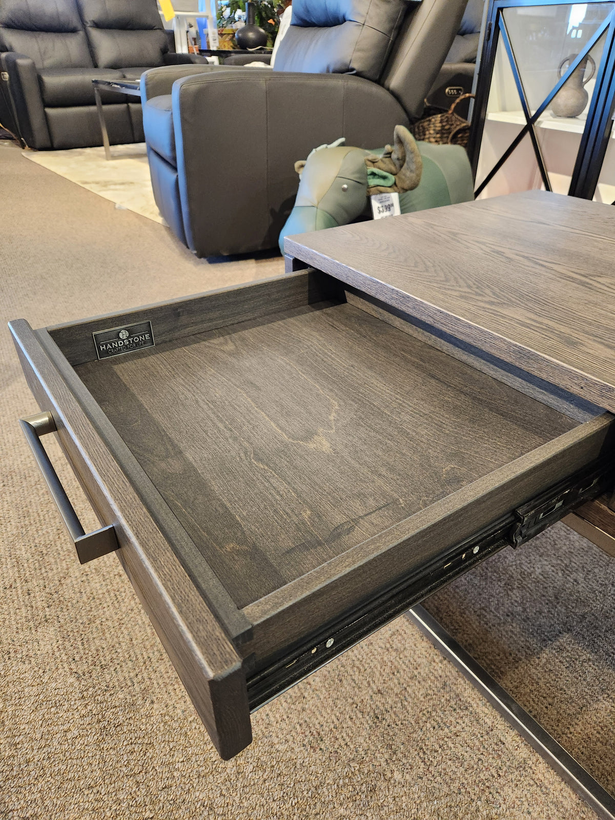 Open the woodgrain drawer of a Muskoka Coffee Table by Handstone, surrounded by a chair and other showroom furniture.