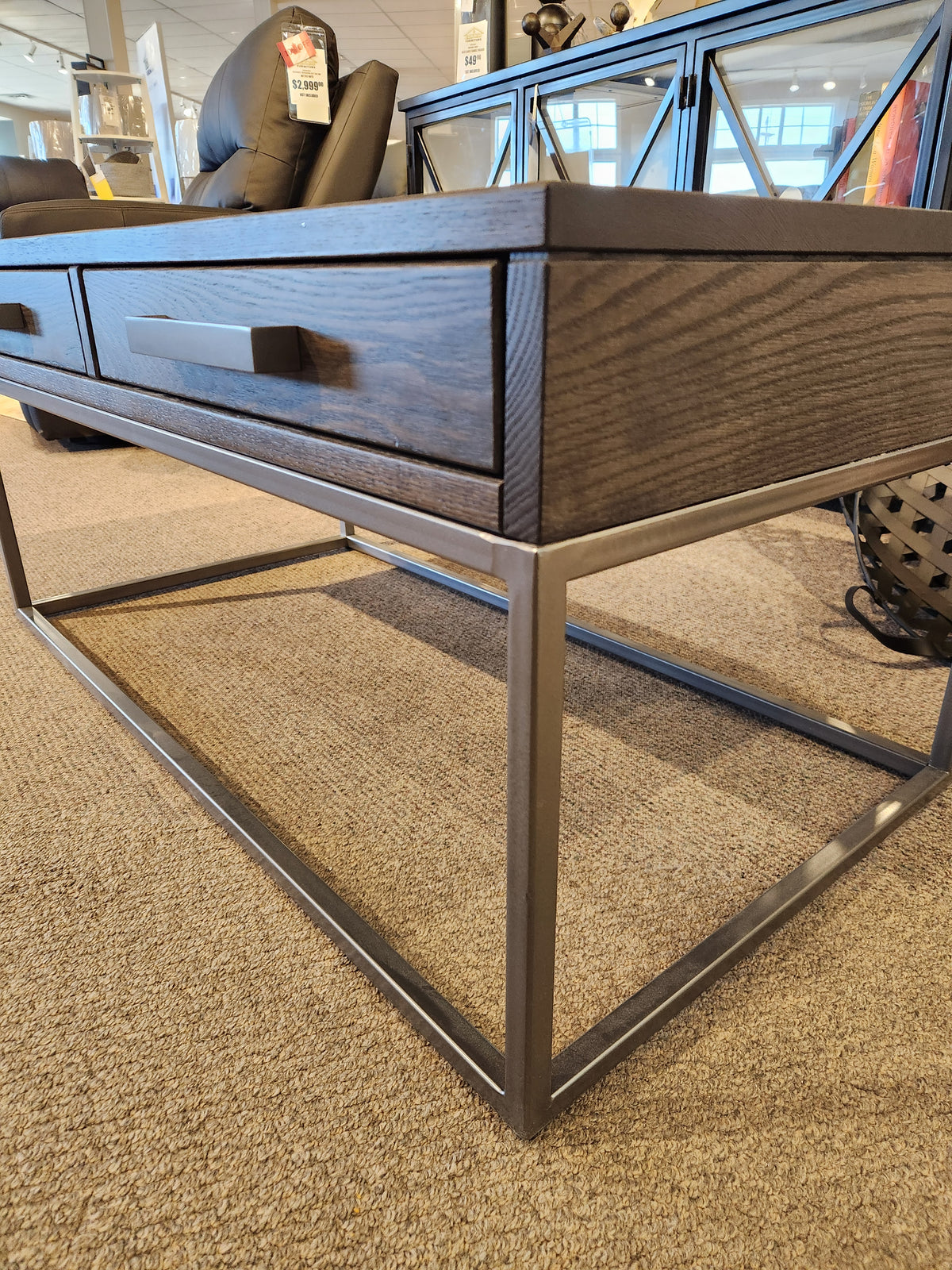 The Muskoka Coffee Table by Handstone features a metal base and drawer, on display in a showroom with price tags nearby.