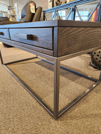 The Muskoka Coffee Table by Handstone features a metal base and drawer, on display in a showroom with price tags nearby.