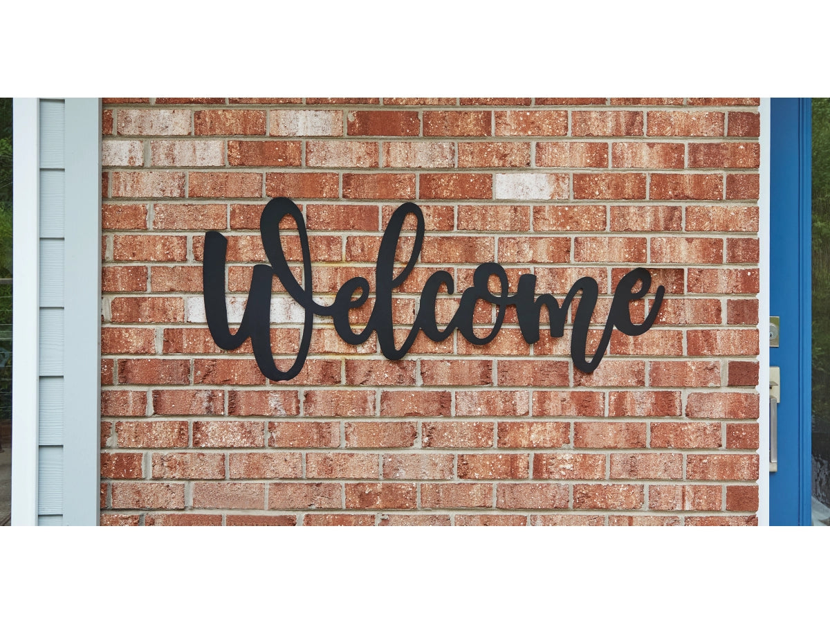 The Emalee Welcome Wall Decor by Ashley, a charming black cursive sign reminiscent of farmhouse style, is gracefully mounted on a red brick wall near the door.