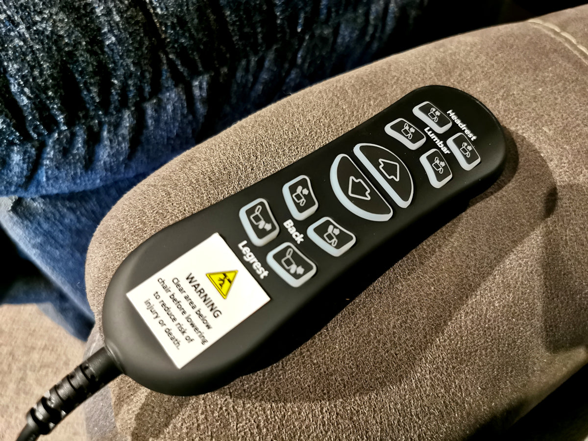 A remote control with numerous buttons and a warning label is positioned on the brown armrest of the Ashley Balliser Lift Chair.