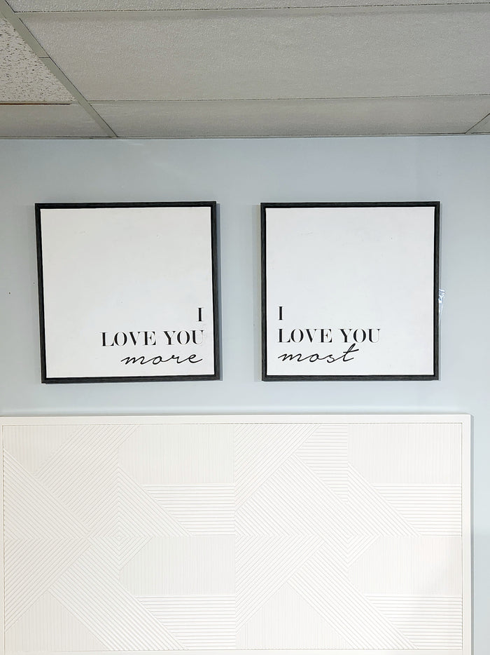 The Adline Wall Art by Ashley features two framed signs set against a white backdrop, with the phrases "I LOVE YOU more" and "I LOVE YOU most" stylishly presented in black font.