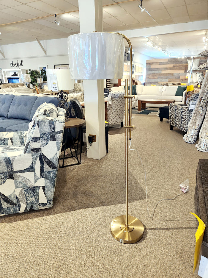 In a furniture store, a Baronvale Floor Lamp by Ashley is showcased with its adjustable arm and sophisticated gold finish, set amidst chic sofas and various home decor accents.