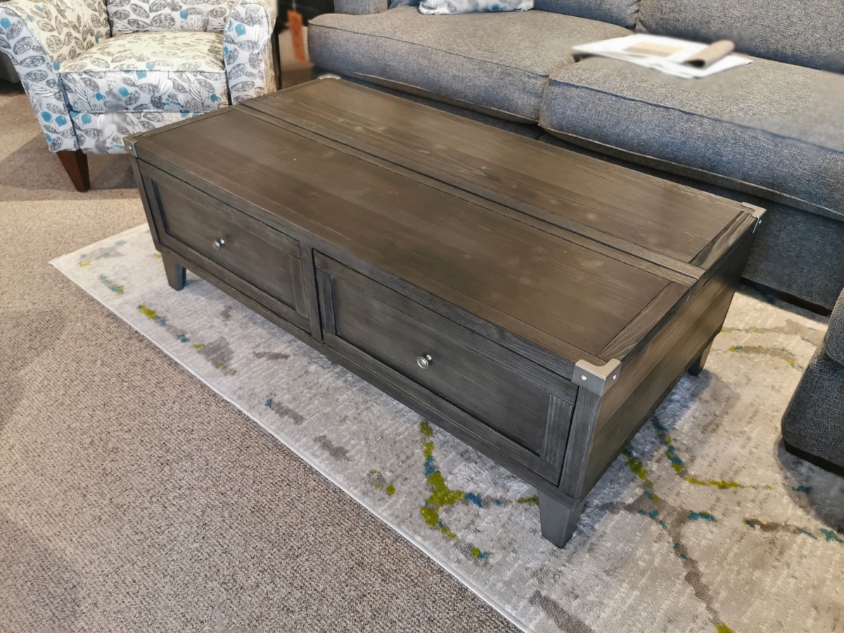 The Ashley Tadoe Lift-Top Coffee Table with two drawers sits on a gray patterned rug in front of a couch and armchair.