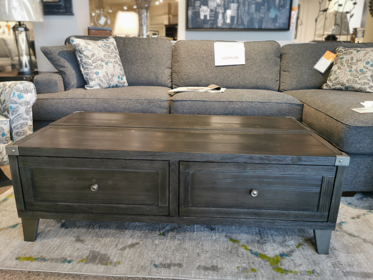 Ashley Tadoe Lift-Top Coffee Table in dark wood with two drawers, positioned in front of a gray sofa with patterned cushions.