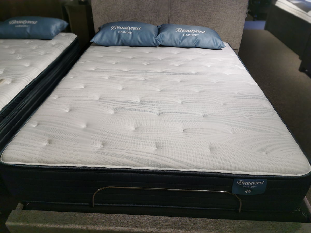 A Beauty-rest Anchorage TT Mattress with a quilted top is displayed on a showroom floor, promising restorative sleep and featuring two blue pillows with branding.