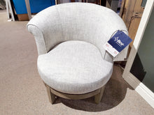 On a carpeted store floor sits the Attica Swivel Chair by Best, designed in a modern light gray with an upholstered cottage style and curved backrest. This comfortable chair can be customized with a variety of fabrics to perfectly suit any space.