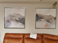 A pair of Crestview Collection CVTOP3322 Galloway Canvas Paintings, featuring earthy tones with gold and silver embellishments, hang above a brown leather couch, where a price tag is visible.